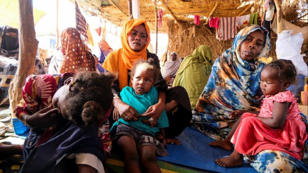 Two women sit holding two young children. An older child sits to the left of them with her face in her hands. They are in a shelter in a displacement camp after fleeing El-Fasher. 