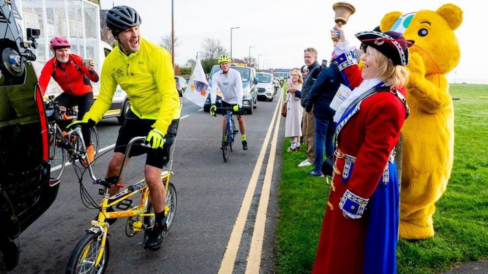 Paddy smiles at a row of people including Pudsey and, Kila Redfearn, a town crier in the seaside town of Lytham St. Anne, where Paddy is cycling through as part of his challenge. 