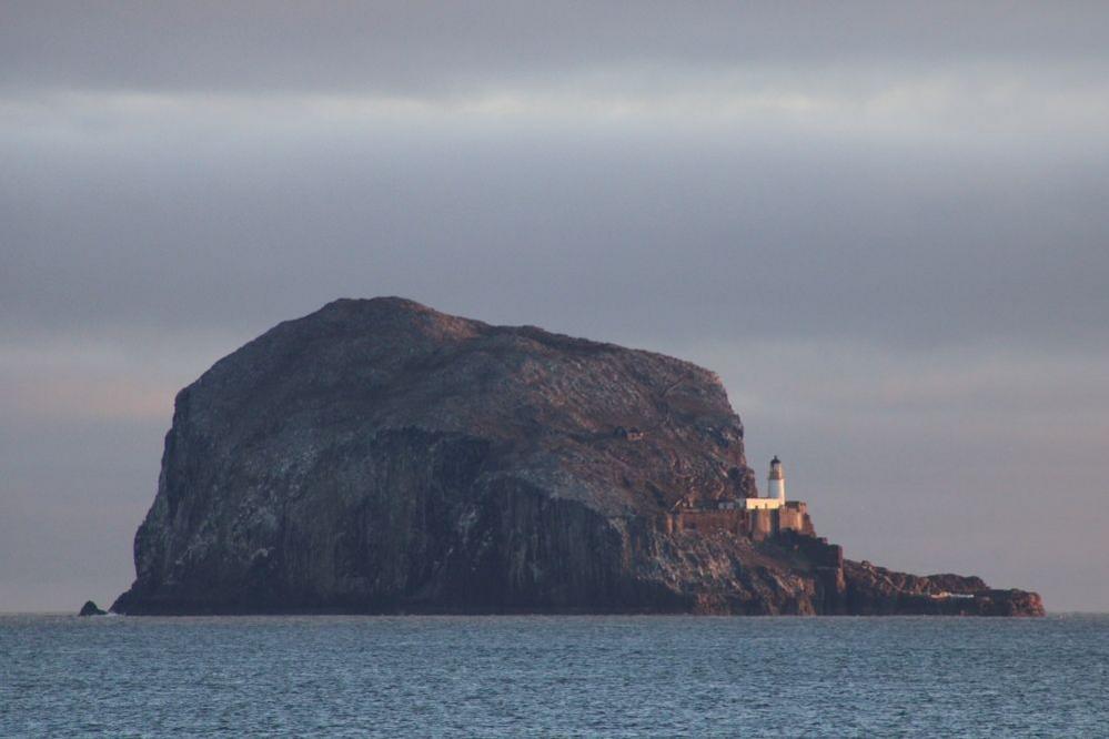 Bass Rock surrounded by water on a cloudy day. A lighthouse stands at the far right side of the rock. The water is calm.