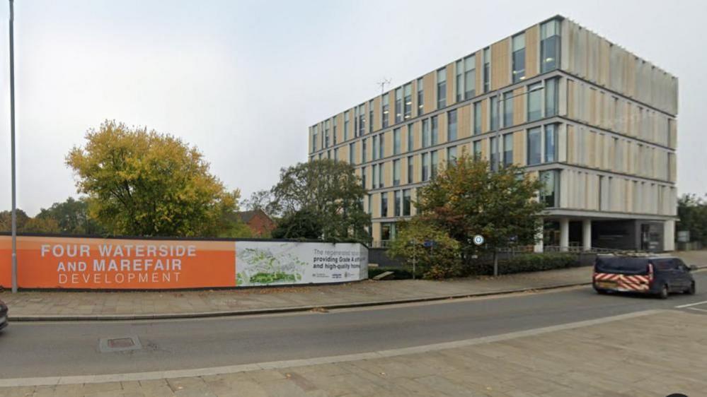 An orange and white banner, with the words "Four Waterside and Marefair", is seen alongside a town centre road. There are trees behind the banner.  To the right is a light brown and grey modern building with five storeys - the University of Northampton Innovation Centre.