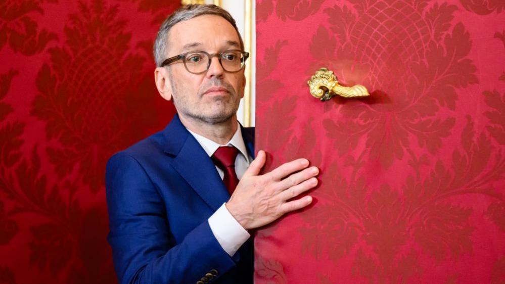 Herbert Kickl, wearing a blue suit jacket and red tie, pushes a red door with a gold handle