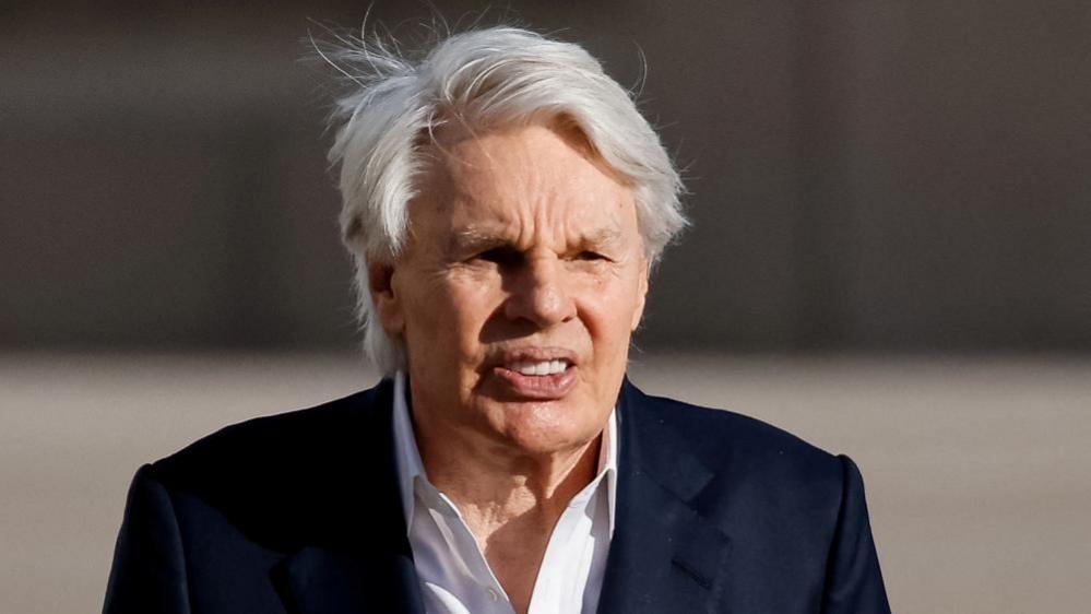 An older man with white hair wears a black suit jacket and a white shirt