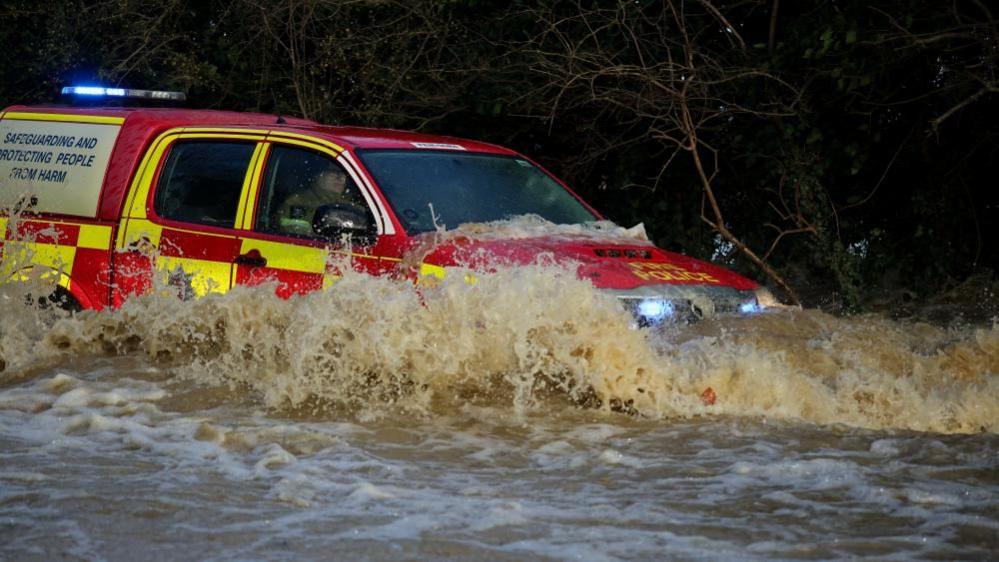 An emergency services vehicles drives through deep water near Billing Aquadrome. It is a 4x4, red and yellow and has its blue lights flashing