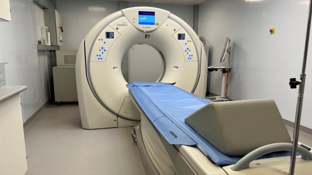 A CT scanner, which is a large circular machine with a blue display screen at the top. A blue cover is on a bed, as is a wedge-shaped pillow. The walls are white.