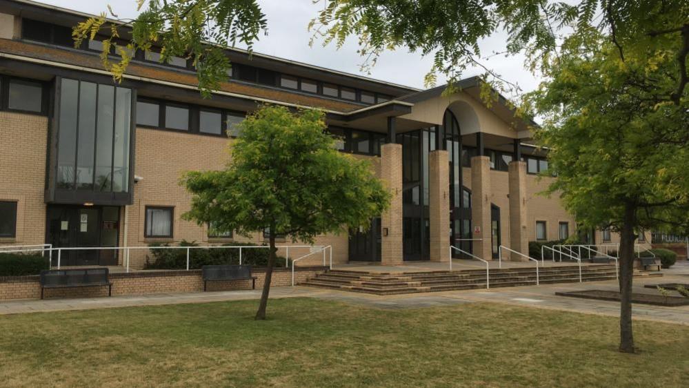 Great Yarmouth Magistrates' Court House, a yellow brick building with brown framed windows. The building has a slate roof and colonnaded facade. There are lawns and trees, as well as pathways and steps on the approach to the entrance.
