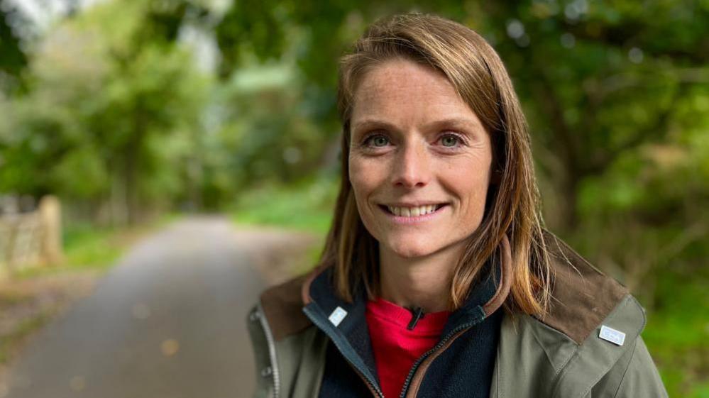 Cath Crowther is standing on a road in a wooded area. She is wearing a green jacket with blue and red tops underneath. She is smiling at the camera.