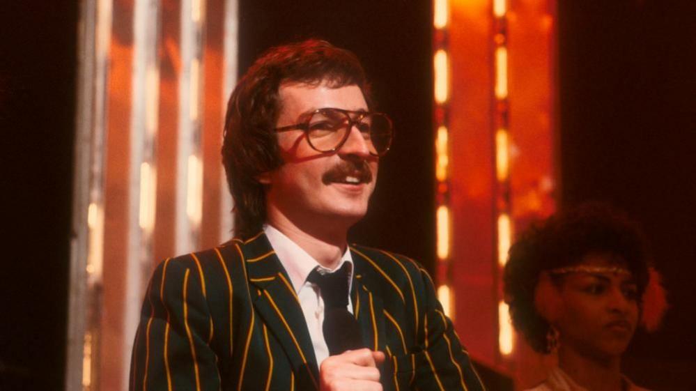 Steve Wright on a set of Top of the Pops, wearing a green jacket with yellow stripes and black tie.