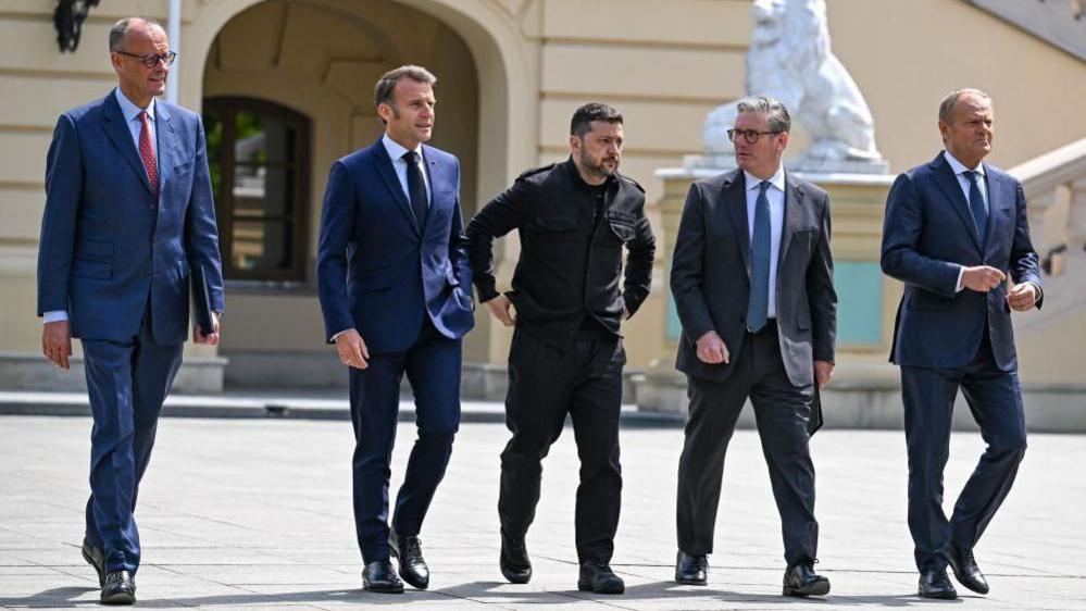 European leaders Merz, Macron, Starmer and Tusk, all wearing suits, walk alongside Zelensky, in dark clothes, across a public square