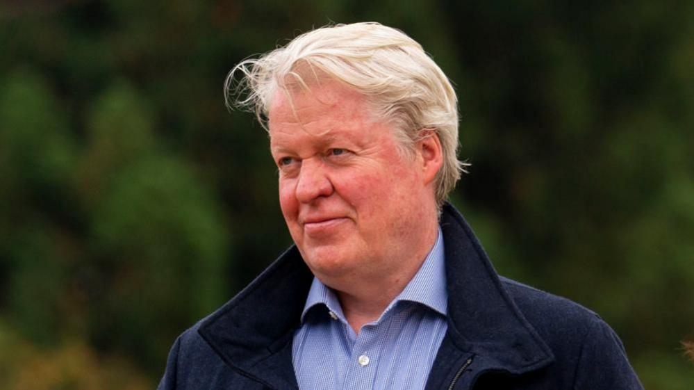 Earl Spencer in a blue shirt and a blue jacket looking to the side of the camera, with blurred trees behind him.