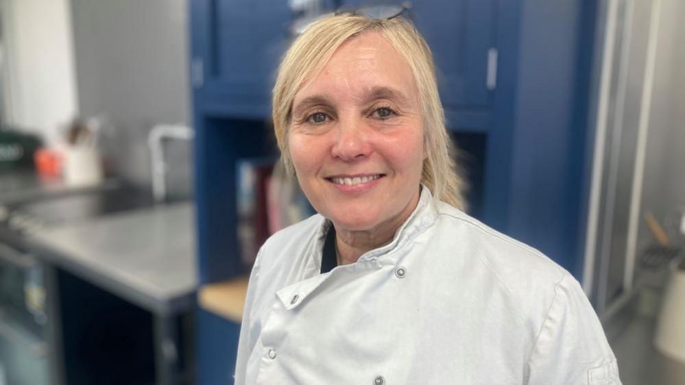 Alison Crouch with long blonde hair, wearing a white chef's jacket, smiling straight at the camera.