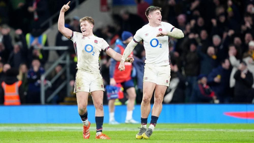 Two Northampton Saints players in white strip with 02 logo on the shirt. One has short ginger hair (Smith) and the other has short dark hair. Fin has his right arm in the air and his left reaching for Freeman's right arm. They are on a grass pitch with spectators behind them.