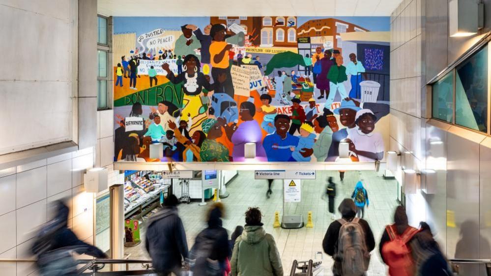Brixton Tube station's entrance with a number of blurred people walking down a staircase. Above the staircase is the bright and colourful mural artwork. At the bottom on the stairs are signs for the Victoria line and two yellow wet floor signs.