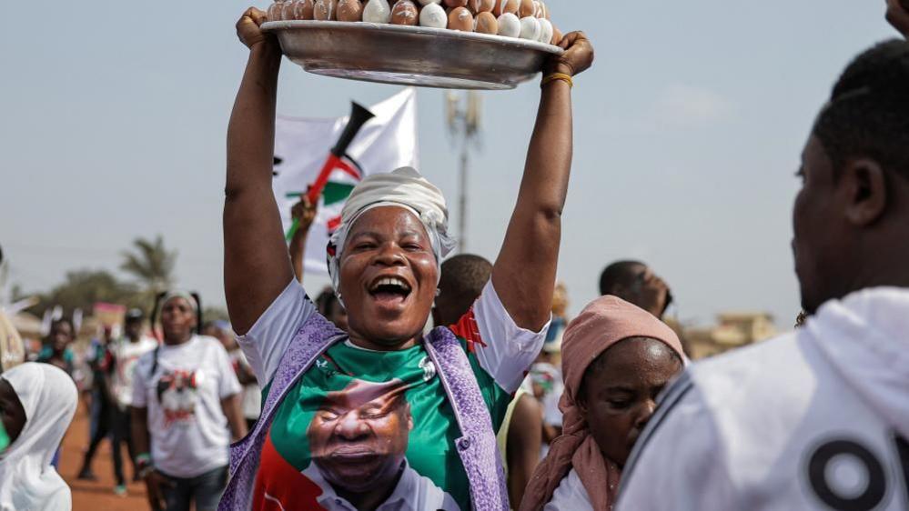 A street vendor, wearing a John Mahama T-shirt, smiles as she hoists a big platter of food above her head, in Accra, Ghana - 5 December 2024