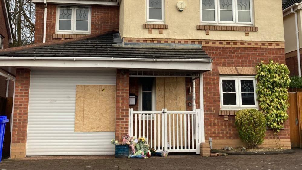 A detached house with the front door and part of the white garage door boarded up with wooden boards. About 10 bunches of flowers are laid near to the front door. 