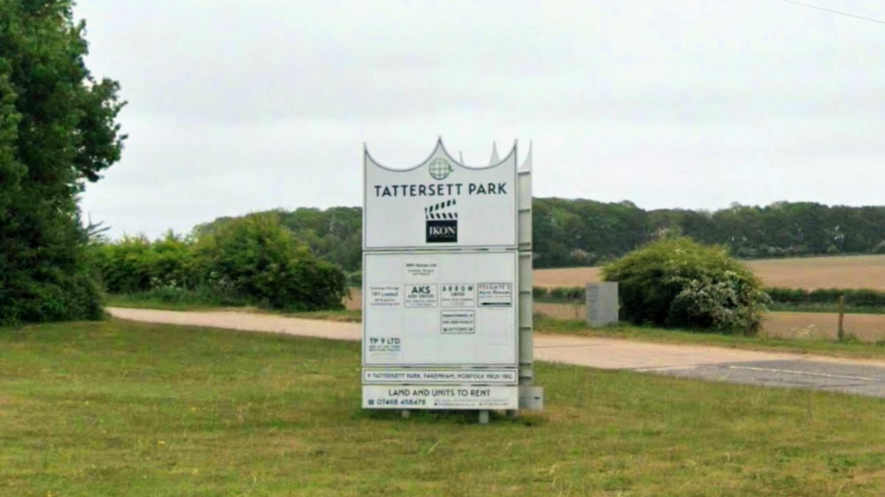 A large white sign welcomes visitors to a business park in a rural loction, with an entrance road alongside it shown with grass and trees on both sides and with a field and woodland in the background.