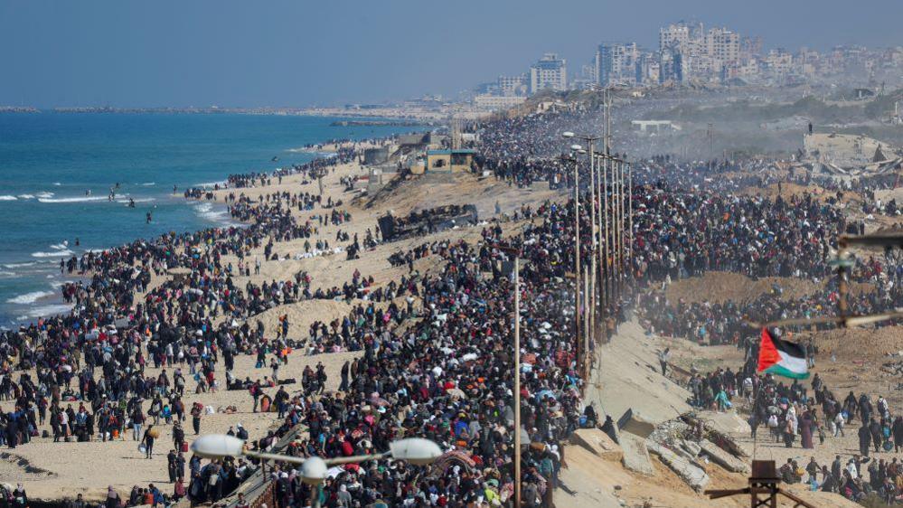 Drone footage shows masses of people walking along a coastal road to return to north Gaza, a city ahead of them and the ocean on their left side, with a Palestinian flag visible 