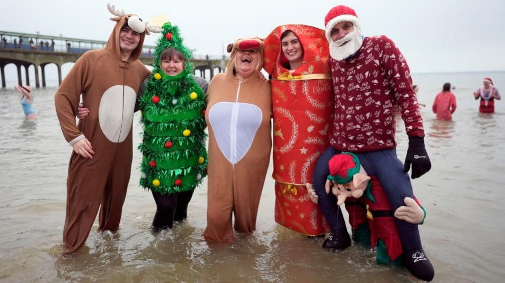 People wearing Christmas fancy dress, including reindeer, a tree and a cracker, stand in six inches of sea water