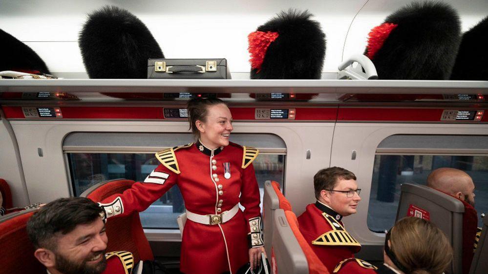 The guards on the train in their uniforms with their hats above them on the luggage racks. One guard is standing up in front of her seat and smiling. Guards around her are chatting and smiling.