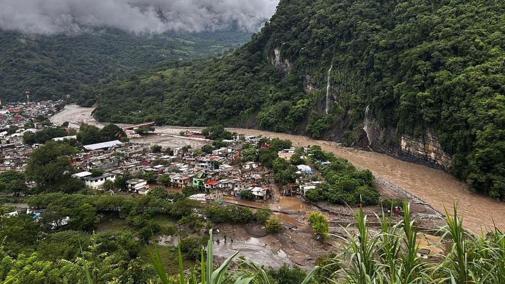 A town is pictured beside a swollen river with lush green trees on either side of the river. The water is brown.