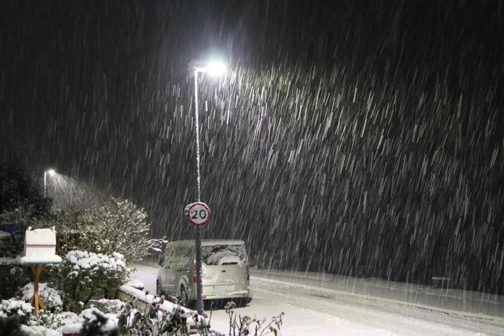 Snow falls in the dark hours of the morning in Lochcarron in the Highlands. A snow covered garden can be seen as well as a car and a 20mph road sign.