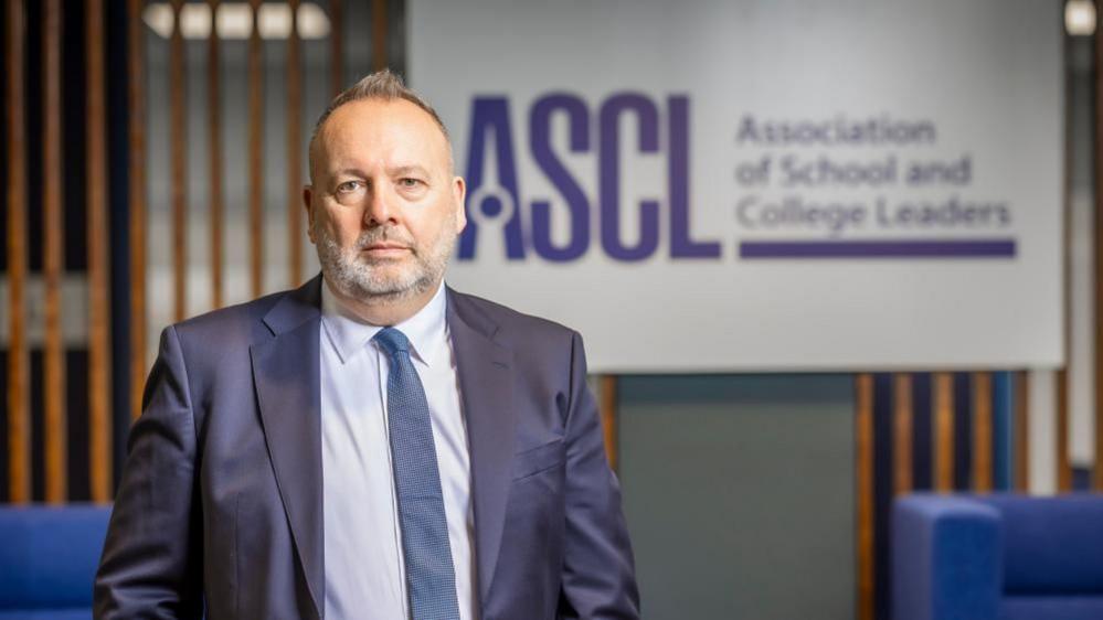 Pepe Di'Iasio, wearing a blue suit, blue tie, and and a light blue shirt. He has a grey beard, receding hair which is light brown and is spiked at the front. He is standing near a wooden screen, with a board stating ASCL, Association of School and College Leaders, and nearby are some blue sofas.