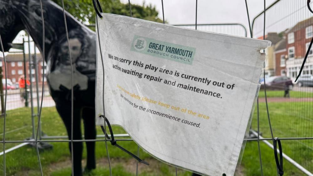 A sign on temporary fencing states: "We are sorry this play area is currently out of action awaiting repair and maintenance. For your own safety please keep out of the area. We apologise for the inconvenience caused." The sign bears the Great Yarmouth Borough Council logo, featuring a shield with three lions heads attached to three herring tails, symbolising the King John Charter of 1208 and the wealth created from the fishing industry.