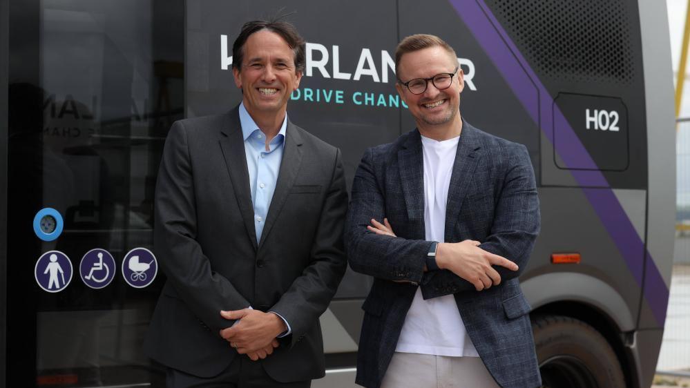 Mike Dawson (left), is standing smiling at the camera. he is wearing a black suit jacket and light blue shirt. He has brown hair. Gavin Jackson, right, is smiling at the camera and has his arms crossed. he is wearing a checked suit jacket and white t-shirt. He is wearing black framed glasses and has blondish hair.