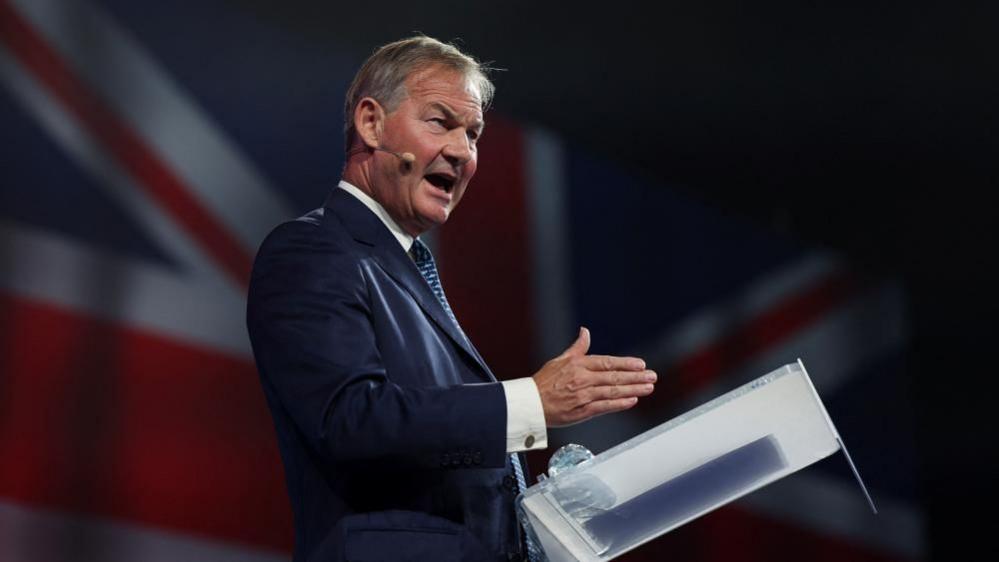 Rupert Lowe looks animated while standing at a lectern on stage, speaking through a hands-free microphone, with an image of a Union Jack behind him. 