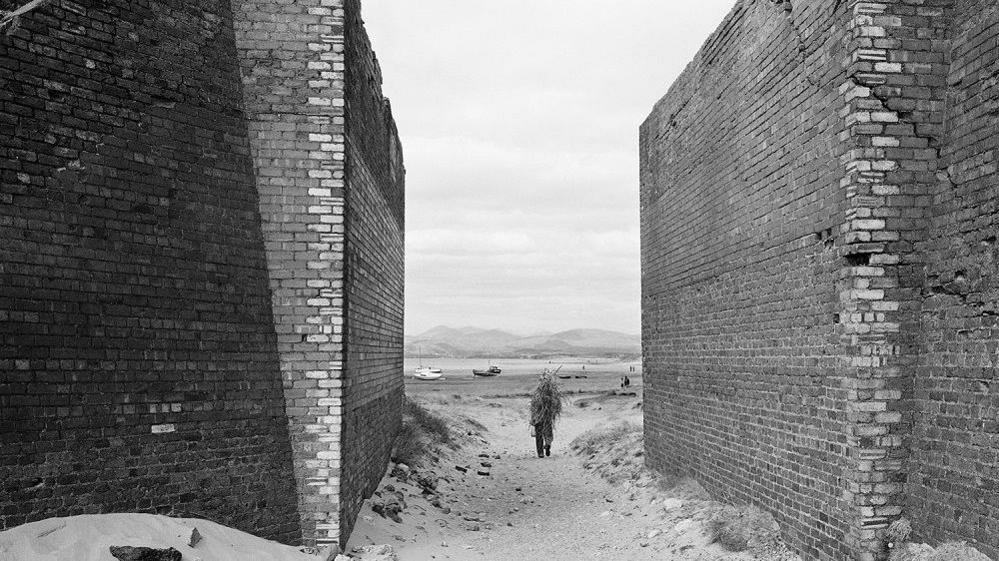 A black-and-white photograph of a man walking on the beach between two large brick walls carrying sticks on his back. The sea, boats and hills can be seen in the distance.