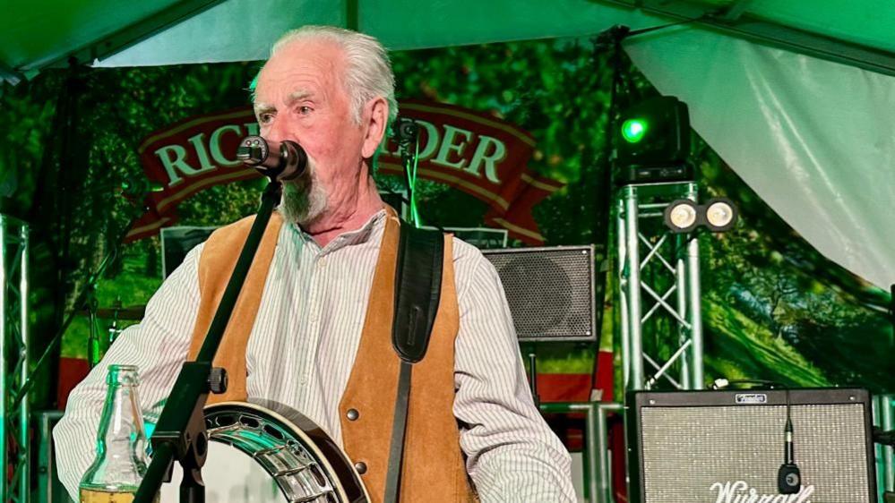 Pete Budd from The Wurzels dressed in a white shirt with brown waistcoat, holding a banjo and singing into a mic. There's stage set and lights behind him inside a green lit marquee