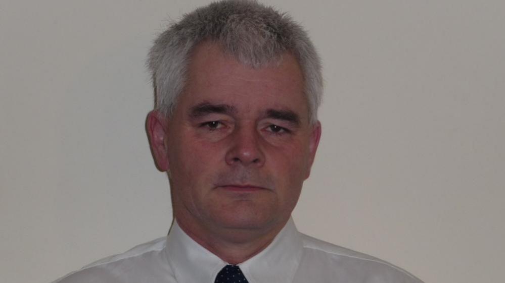 It is a head shot of Geoff Lynch. The background is a cream wall and Mr Lynch is wearing a white shirt with black tie. 