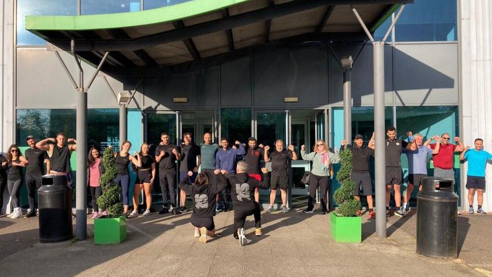 People stand in a line flexing their biceps in front of the Nuffield Health gym. Two people kneel in front of them with their backs to the camera showing their hoodie which reads 25.