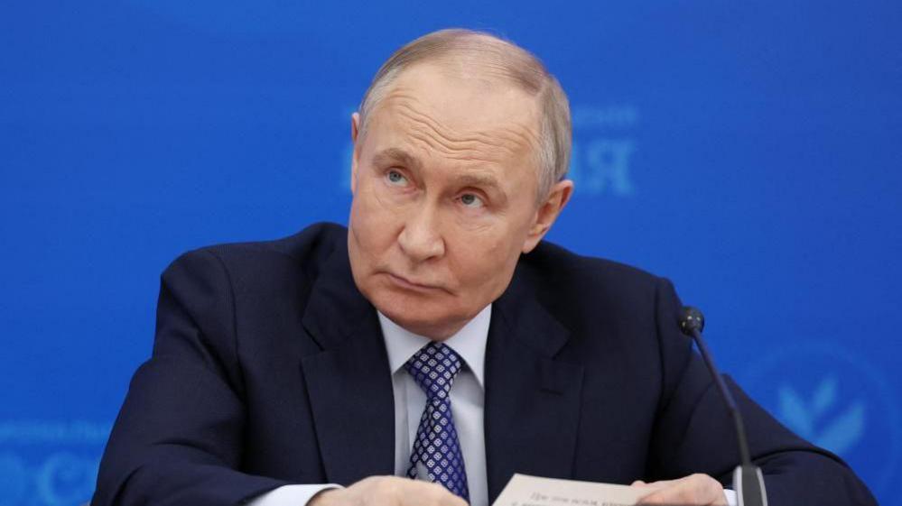 Russian President Vladimir Putin sits in front of a microphone, wearing a dark suit and navy tie, during a meeting in Moscow.