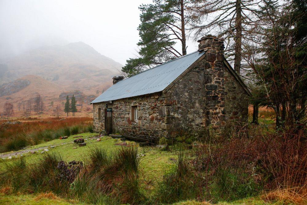 Corryhully bothy, a stone house with two chimneys. A hill in the background is obscured by a light fog.