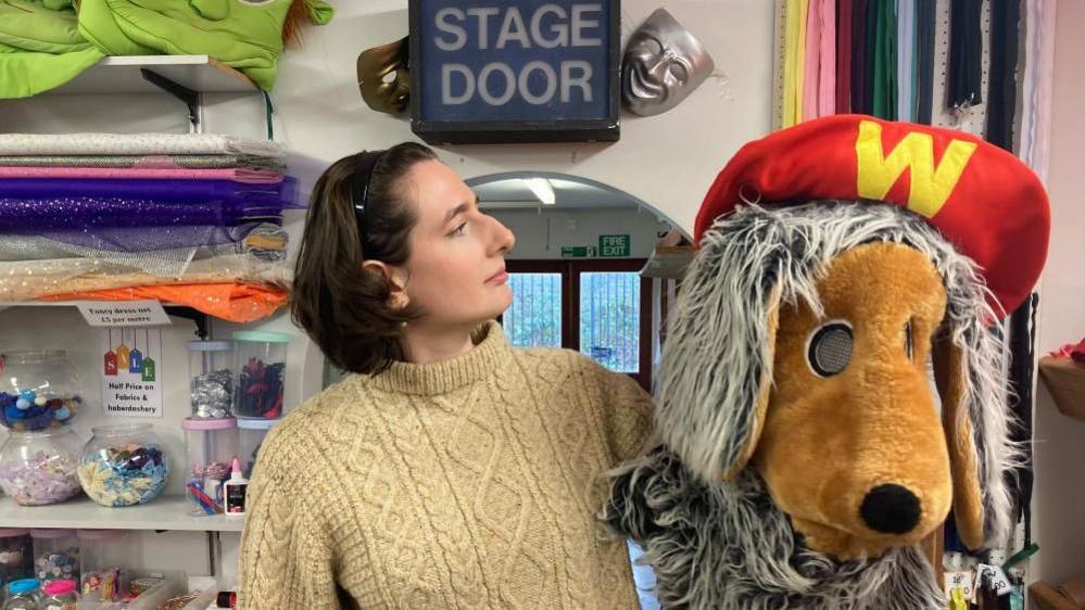 A dark-haired woman wearing a cream round-necked jumper looks at the head of a Womble costume she is holding in her left hand. Above her head is a sign saying "stage door".