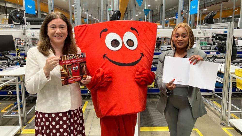 Alex Mayer, holding a book, wears a white jacket and red and white spotted dress. There is a red cartoon character in the middle and Sally Kimondo, wearing grey leggings and a grey jacket, is next to it. 
