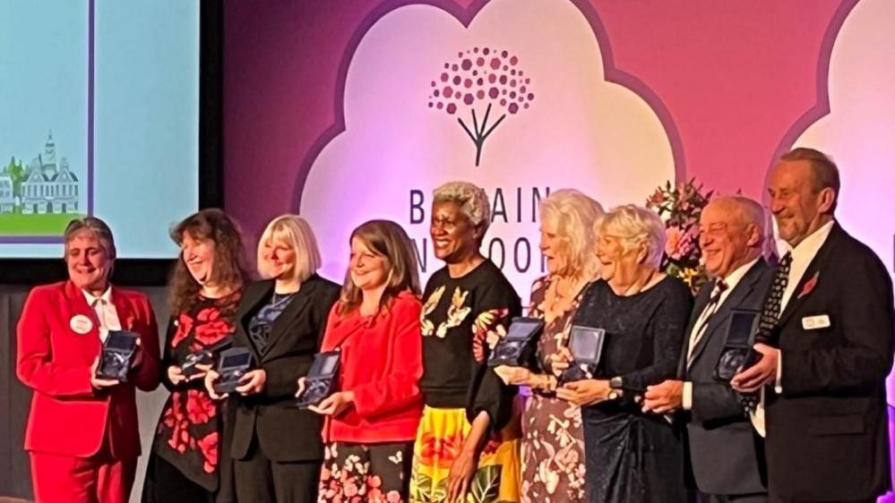 A group of women and men lined up on a stage, holding awards. Behind them is a backdrop promoting RHS Britain in Bloom.
