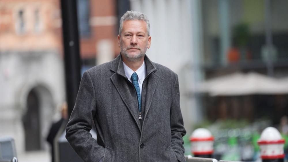 Nathan Gill wears a grey coat with a white shirt and blue tie underneath. He has his hands in his pocket and looks away from the camera. He has short grey hair and grey stubble. 