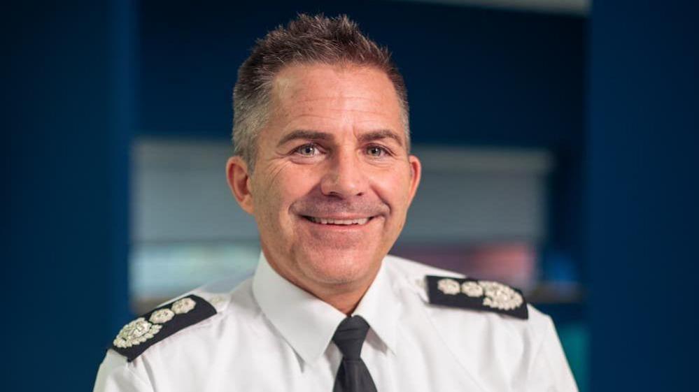 A Simon Tuhill pictured smiling with short dark spikey hair. He is wearing a white shirt and black tie with black Epaulettes on his shoulders.