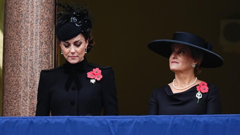 Catherine, dressed in black, looks down. Next to her, Sophie, also in black, looks up to the left.