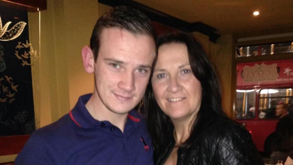 Josh Hanson with short dark hair smiling at the camera and wearing a blue polo shirt with red trim. He is standing very close to his mother, who has long dark hair and is wearing a black leather jacket. They are in a house with wooden shelves behind.