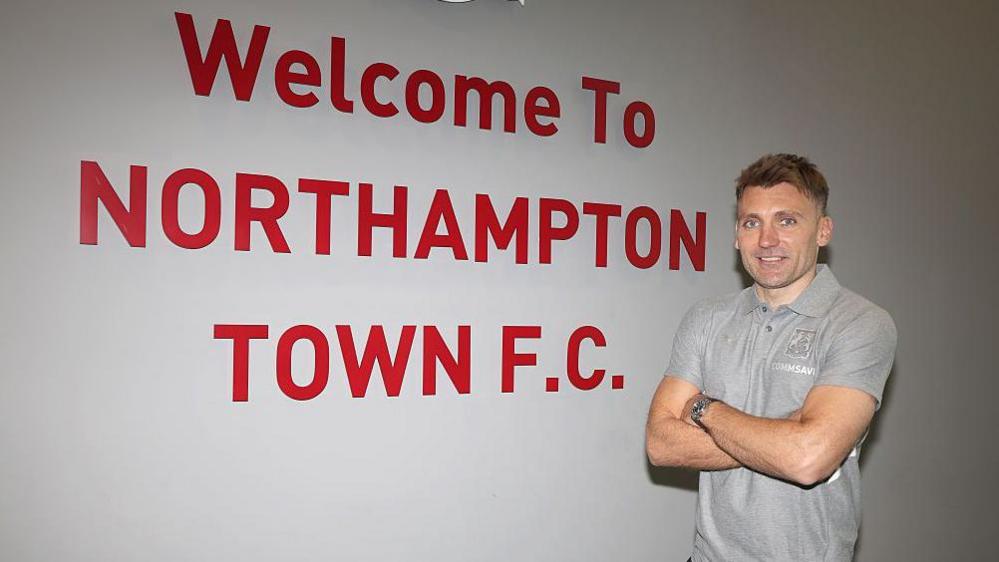 New Northampton Town manager Chris Hogg with his arms folded in a grey t-shirt standing in front of a white wall with Northampton Town FC written in red letters on it
