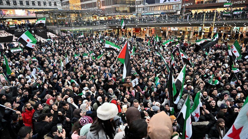 Very large crowd of people waving Syrian flags in  Sergel's Square in Stockholm, Sweden December 8, 2024
Raised buildings and walkway barely visible in the distance