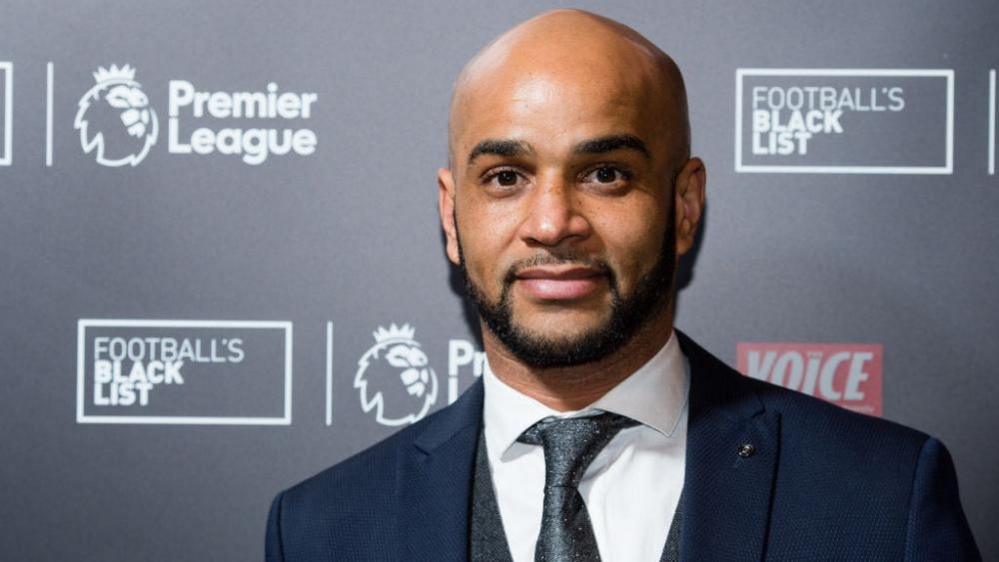 Leon McKenzie is dressed in a white shirt, grey tie, grey waistcoat and dark blue jacket. He has a shaved head and a trimmed beard and moustache. He is standing in front of Premier League branded hoarding, with other imagery including a statement that reads: "Football's Black List".