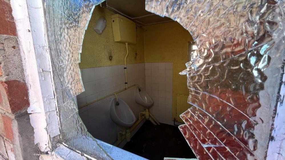 A view of the men's toilet urinals viewed through the broken obscured glass of the Crittall window. The image shows tiled panelling on the wall, with yellow paintwork and a cistern painted yellow, with broken glass held within a frame, cracked, and a large opening through the glass.