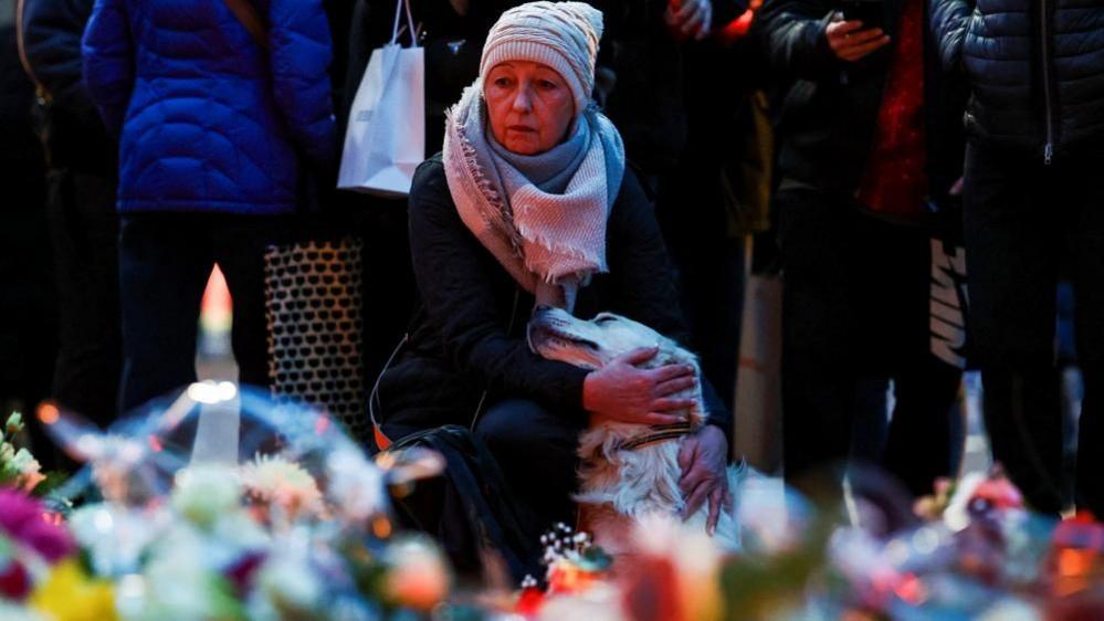 A woman pets a dog as people leave floral tributes to the victims near the site where a car rammed into a crowd at a Magdeburg Christmas market in Magdeburg, Germany December 21, 2024. The woman is wearing a white hat and scarf. 