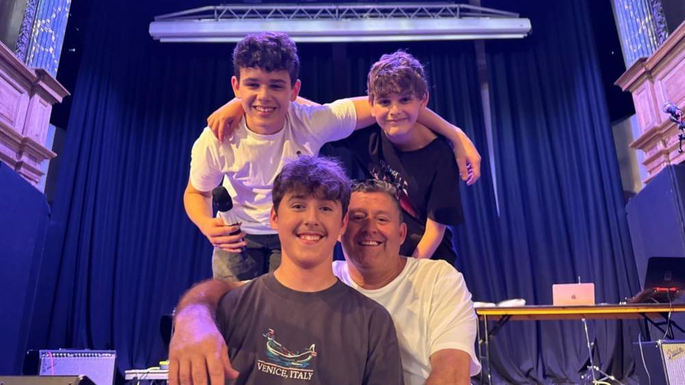 Dan and his dad Shaun sitting on the stage. Dan is wearing a brown tee shirt with a logo. Shaun is wearing a white tee shirt, and has his right arm draped over his son's shoulder. Behind them, with arms around each other, are two other band members. They are on the stage in St George's Theatre with curtains behind and a gantry rig above them..