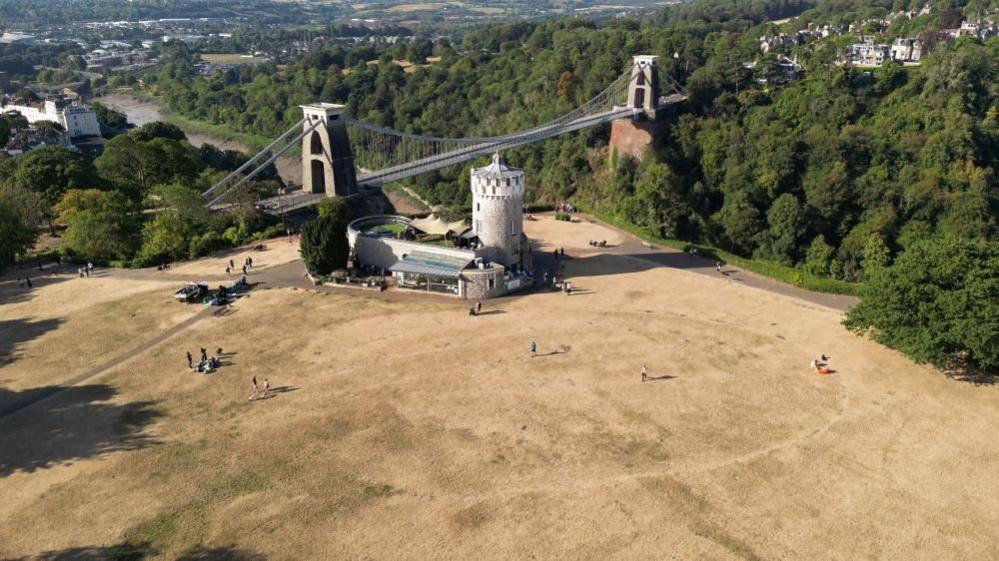 An aerial view of Clifton Observatory in Bristol surrounded by brown, dry ground. In the background is the Suspension Bridge and trees lining the gorge.