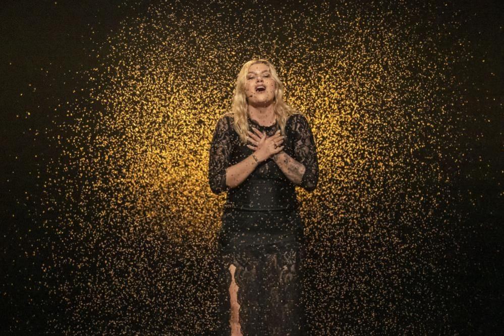 France's Eurovision entry Louane crosses her hands across her chest as she sings with her eyes closed, wearing a black dress and surrounded by a gold confetti-like effect, with a black backdrop.