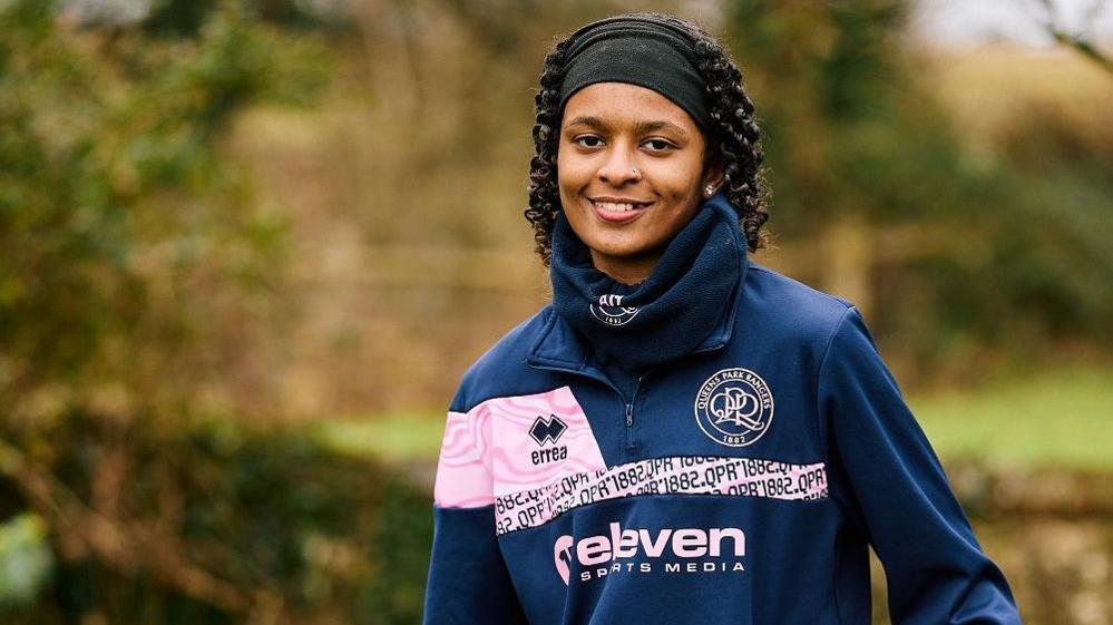 Amani wearing a pink and blue QPR training top and snood. She has her hair pulled back by a black headband and is smiling at the camera. 